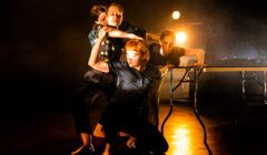 Four dancers in intense, dynamic poses on a dark stage with dramatic lighting; one dancer is knelt over while the others support and interact around her. A table is visible on the right side of the stage.