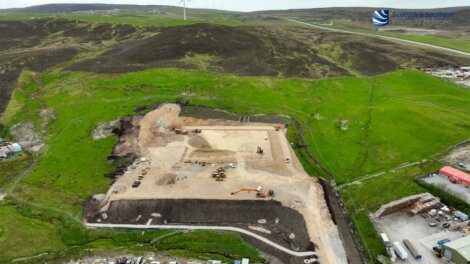 Aerial view of a construction site in a hilly green area with wind turbines in the background, heavy machinery and earth-moving activities are visible at the center of the site.