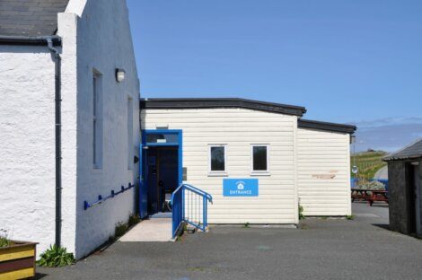 Entrance to a white and light grey building with a blue handrail and door, marked by a blue sign with the word "Entrance." The area is paved, and the sky is clear.