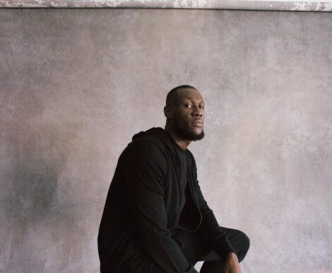 A man in a black hoodie sits against a mottled gray backdrop, looking towards the camera.