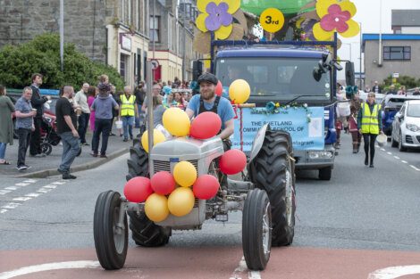 A man drives a decorated tractor with red and yellow balloons in a parade. A truck with colorful flowers follows behind, and people walk alongside and watch from the street.