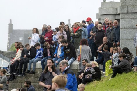 A group of people of various ages sit and stand on stone steps in an outdoor setting, some looking into the distance, while others engage with each other.