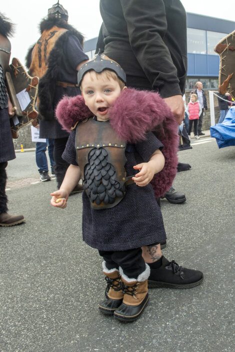 A young child dressed in a Viking costume, including helmet, armor, and boots, stands on a street during an outdoor event with other adults and children present.