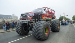 A red monster truck with large tires drives down a road during a parade with spectators on both sides.