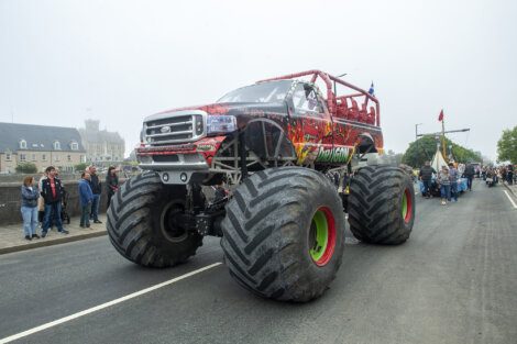 A red monster truck with large tires drives down a road during a parade with spectators on both sides.