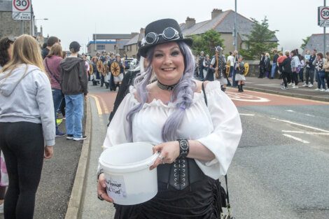 A woman dressed in a steampunk outfit holds a white bucket during a street event. People in the background are gathered on the sidewalk.