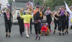 A group of people in colorful outfits and leis happily marching and dancing on a street during a parade. A truck is visible in the background.