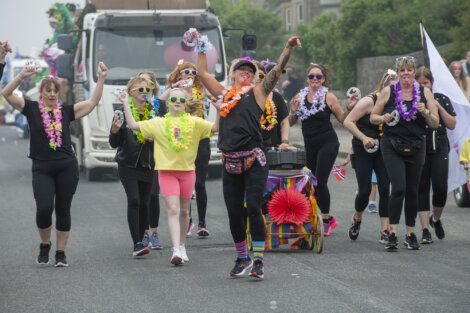 A group of people in colorful outfits and leis happily marching and dancing on a street during a parade. A truck is visible in the background.