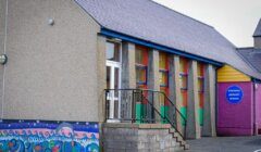 Exterior of a primary school building with colorful murals and a sign reading "Tingwall Primary School" on a pink wall. The building has a gray roof and multiple windows with green, red, and yellow frames.