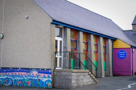 Exterior of a primary school building with colorful murals and a sign reading "Tingwall Primary School" on a pink wall. The building has a gray roof and multiple windows with green, red, and yellow frames.