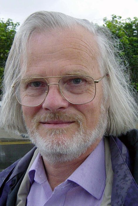 A man with shoulder-length gray hair, glasses, and a beard, wearing a purple shirt and a dark jacket, standing outdoors.