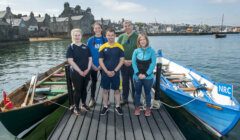 A group of six people stand on a wooden dock in front of two small boats labeled NRC, with a coastal town in the background.