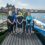 A group of six people stand on a wooden dock in front of two small boats labeled NRC, with a coastal town in the background.