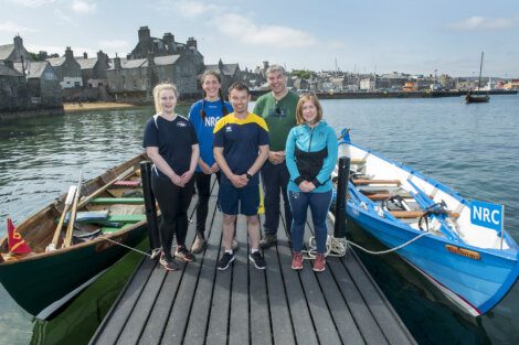 A group of six people stand on a wooden dock in front of two small boats labeled NRC, with a coastal town in the background.