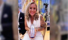 A woman with long blonde hair, wearing a white dress, is smiling and holding a crystal award while seated at an event.