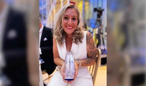 A woman with long blonde hair, wearing a white dress, is smiling and holding a crystal award while seated at an event.