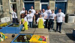 A group of people in matching white t-shirts and gloves are gardening in blue planters outside a building. Some bags of soil and supplies are on the wooden benches.