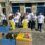 A group of people in matching white t-shirts and gloves are gardening in blue planters outside a building. Some bags of soil and supplies are on the wooden benches.