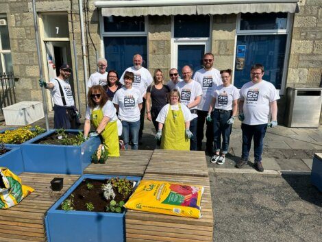 A group of people in matching white t-shirts and gloves are gardening in blue planters outside a building. Some bags of soil and supplies are on the wooden benches.