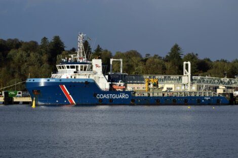 A large blue coastguard vessel docked at a pier with a tree-covered hillside in the background. The ship has a white and red striped emblem and various equipment on deck.