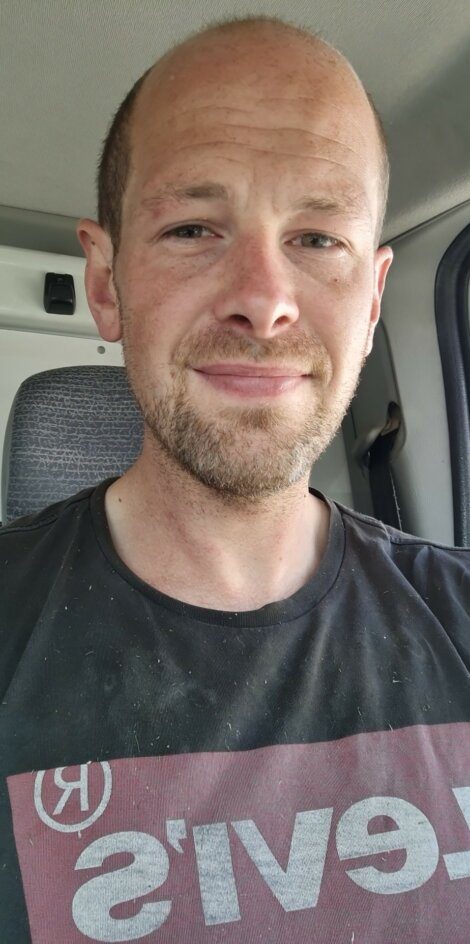 A man with a slight smile, wearing a black T-shirt with a partial logo, sits in a vehicle.