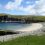 A small harbor with a dock and several boats is set against green hilly terrain. There is a sandy beach and a rocky shore in the foreground under a partly cloudy sky.