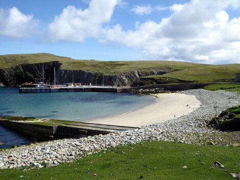 A small harbor with a dock and several boats is set against green hilly terrain. There is a sandy beach and a rocky shore in the foreground under a partly cloudy sky.