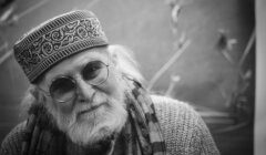 Black and white photo of an elderly man wearing round glasses, a patterned cap, and a scarf. He has a white beard and mustache and is looking at the camera.