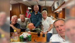 Five people smiling and posing together in a cozy boat cabin with a Norwegian flag in the background. One person holds a decorated pumpkin. Drinks and snacks are on the table.