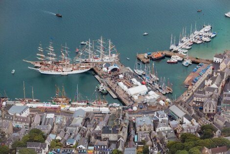 Aerial view of a busy harbor filled with tall ships and other boats, surrounded by a coastal town. Multiple docks extend into the water, with buildings and streets visible in the foreground.