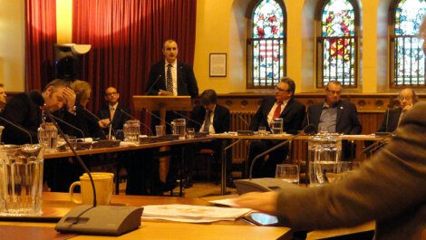 A man speaks at a podium in a conference room with several people seated around tables. Three large stained glass windows are in the background.