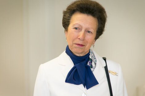 A woman with short, styled hair wears a white blazer, blue scarf, and a pin. She has a thoughtful expression and stands before a neutral background.