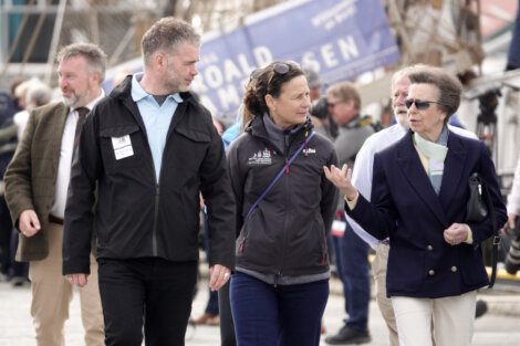 Three people are walking outdoors, engaged in conversation, with a nautical environment in the background.