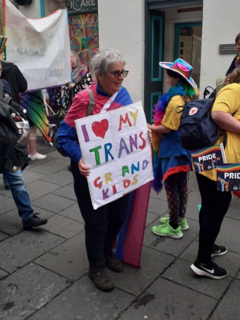 An older person in a festive outfit holds a sign that reads, "I love my trans grandkids," at a Pride event. They are surrounded by others wearing colorful attire.