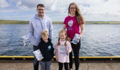 Two adults and two children posing on a dock by the water, holding booklets. The adults wear hoodies, the woman also wears a pink T-shirt, and the children wear a navy hoodie and a light pink T-shirt.