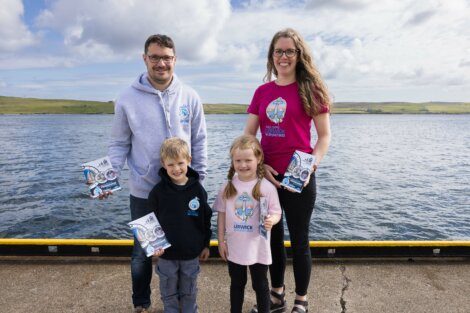 Two adults and two children posing on a dock by the water, holding booklets. The adults wear hoodies, the woman also wears a pink T-shirt, and the children wear a navy hoodie and a light pink T-shirt.