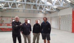 Four people stand inside a partially renovated building with an exposed ceiling and construction equipment in the background.