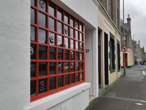 A street view showing the exterior of building number 33 with a large window featuring red grid framing and artworks of faces. Other buildings and part of the road with a no entry sign are visible.