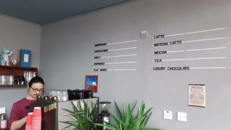 A barista stands behind a counter preparing a drink, with a menu of coffee and other beverages displayed on the wall to the right. Two potted plants are in the foreground.