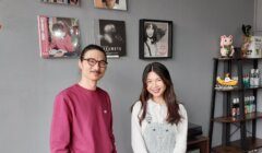 A man in a pink sweater with glasses and a woman in a white top and overalls stand in front of a wall displaying music album covers. Shelving units with various items are visible to their right.