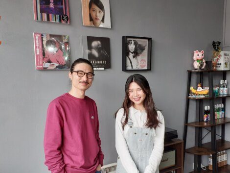 A man in a pink sweater with glasses and a woman in a white top and overalls stand in front of a wall displaying music album covers. Shelving units with various items are visible to their right.