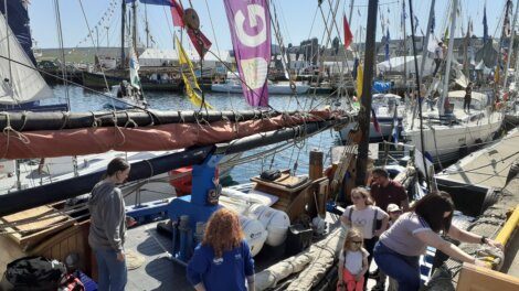 People stand and interact on and around a docked sailboat at a marina, with colorful flags and other boats in the background on a sunny day.