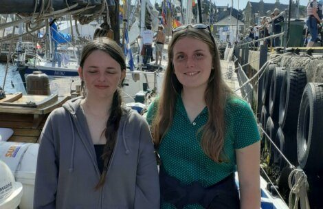 Two young women stand on a dock beside a marina. One has her eyes closed and is wearing a gray hoodie, and the other is smiling while wearing a green polka dot shirt. Sailboats and flags are visible in the background.