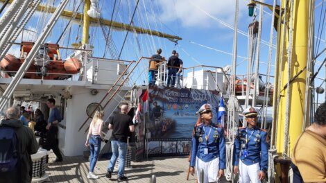 A group of people walk and stand on the deck of a ship with a banner reading "Welcome Aboard." Two officers in uniform are seen in the foreground.