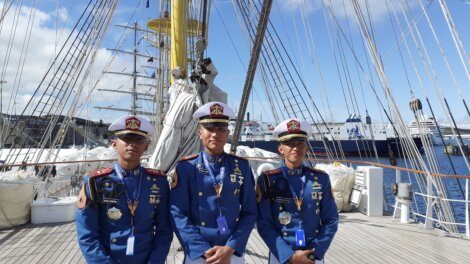 Three uniformed cadets stand on the deck of a tall ship, with sails and rigging visible behind them, against a backdrop of a harbor with other ships.