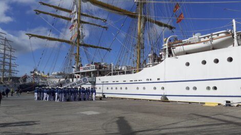 A group of uniformed individuals stands in formation on a dock beside a large white sailing ship with tall masts and rigging. The ship displays a banner reading "INDONESIA.