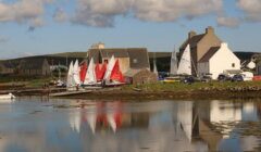 Small sailboats with colorful sails are docked near rustic buildings by a calm waterfront under a cloudy sky.