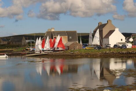 Small sailboats with colorful sails are docked near rustic buildings by a calm waterfront under a cloudy sky.