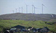 A series of wind turbines on a grassy hill with several houses and buildings in the foreground.