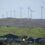 A series of wind turbines on a grassy hill with several houses and buildings in the foreground.